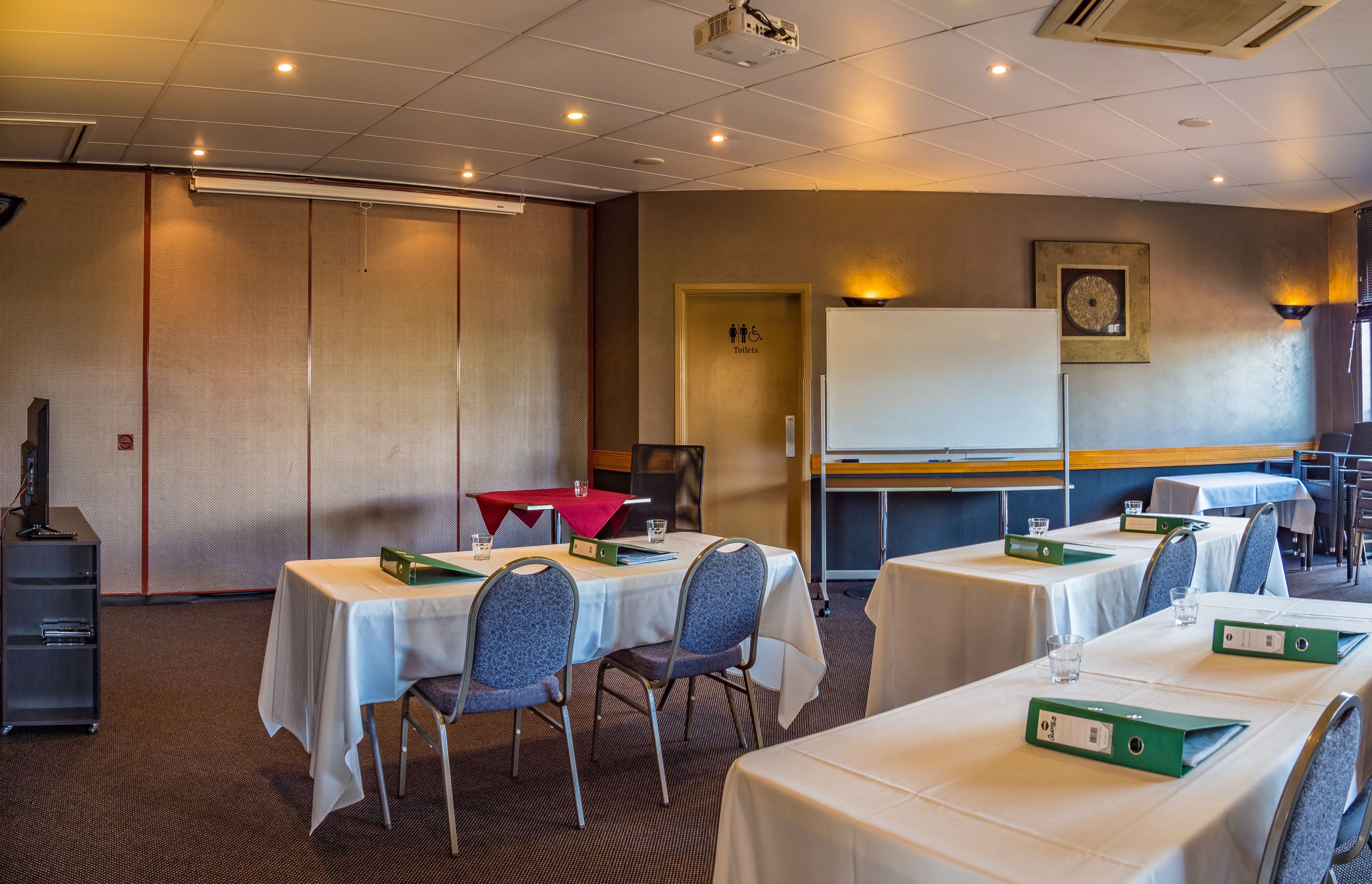 A conference room with tables, chairs, a projector, a whiteboard, and a door labeled "Toilets".