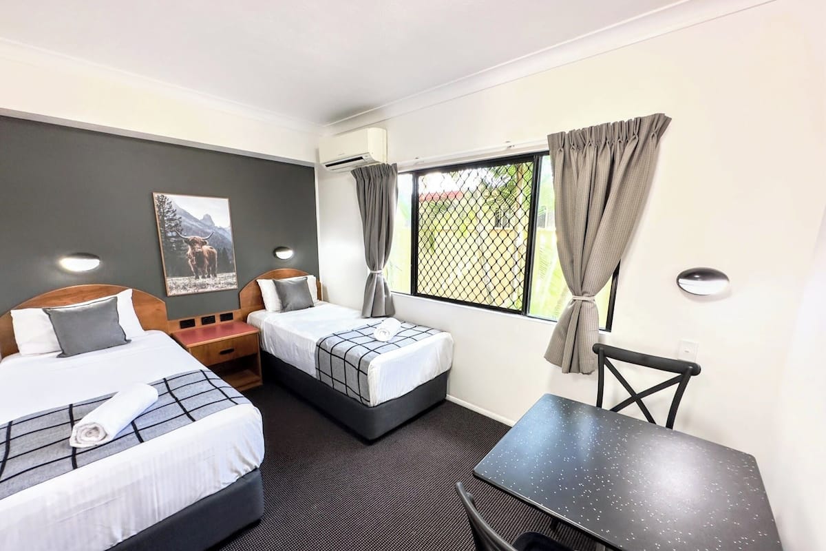 A hotel room with two beds, a desk, and a chair. There is a window with curtains, an air conditioning unit, and a wall-mounted light.