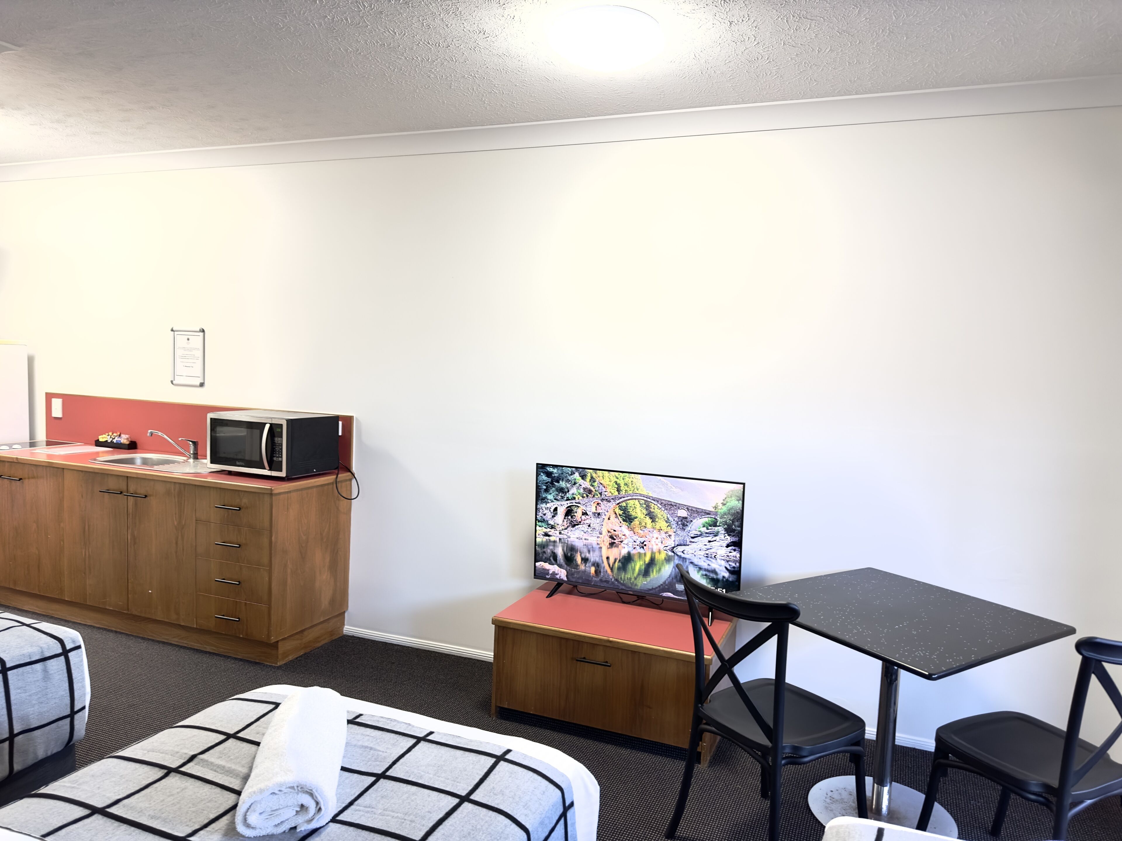 A compact hotel room with a kitchenette, a flat-screen TV, a small dining table, and a sofa with a rolled-up towel.