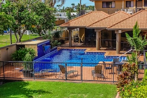Una piscina con sillones de descanso, un porche cubierto y un área cercada con vegetación.