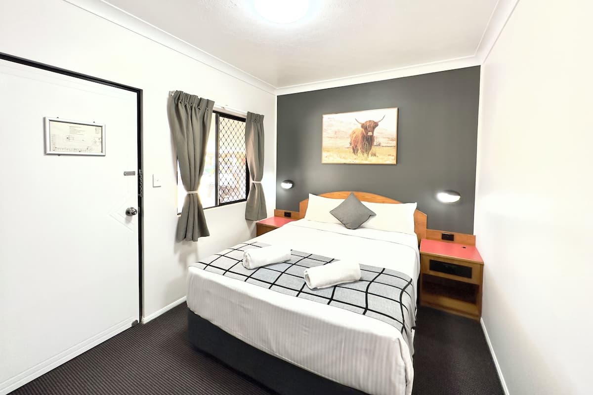 A hotel room with a bed, a nightstand, a wall-mounted picture, and a door with a security camera.