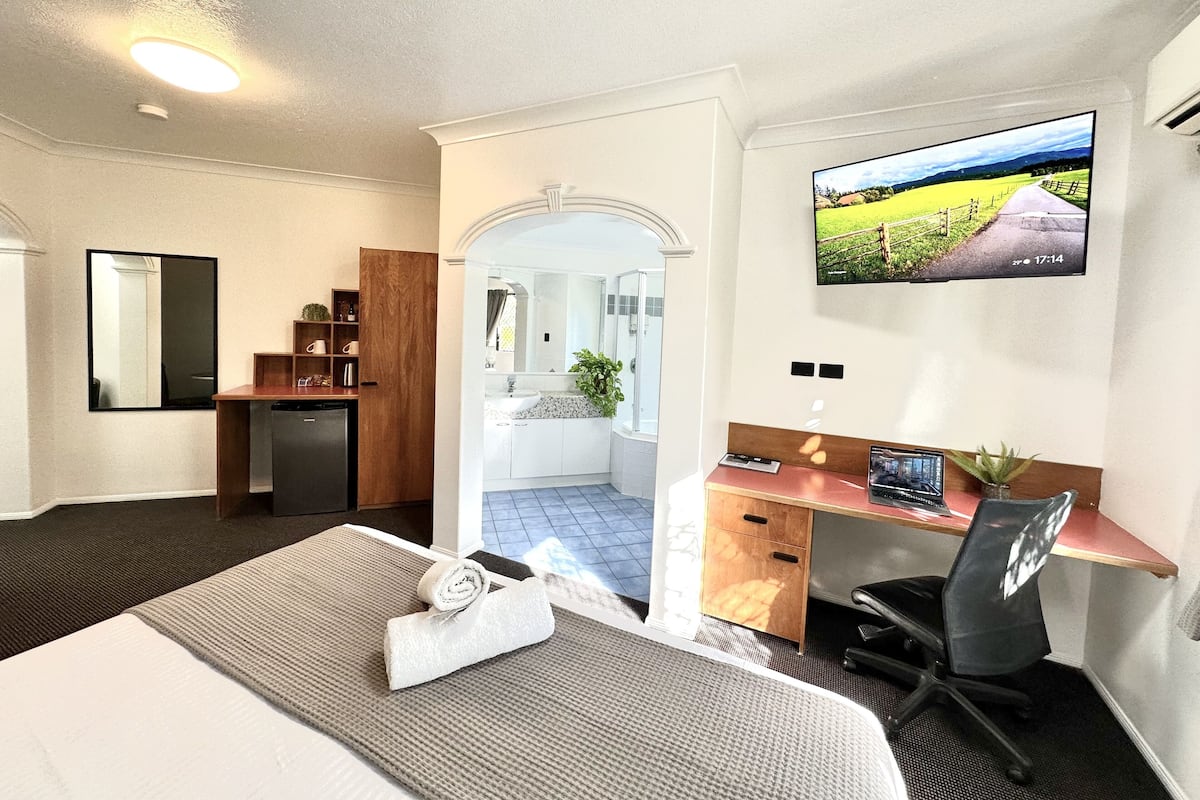 A hotel room with a bed, a desk, a chair, a TV, and a bathroom visible through an archway.