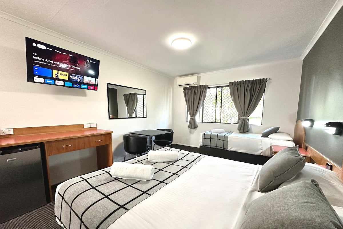 A hotel room with a bed, a desk, a chair, a TV mounted on the wall, and a window with curtains.