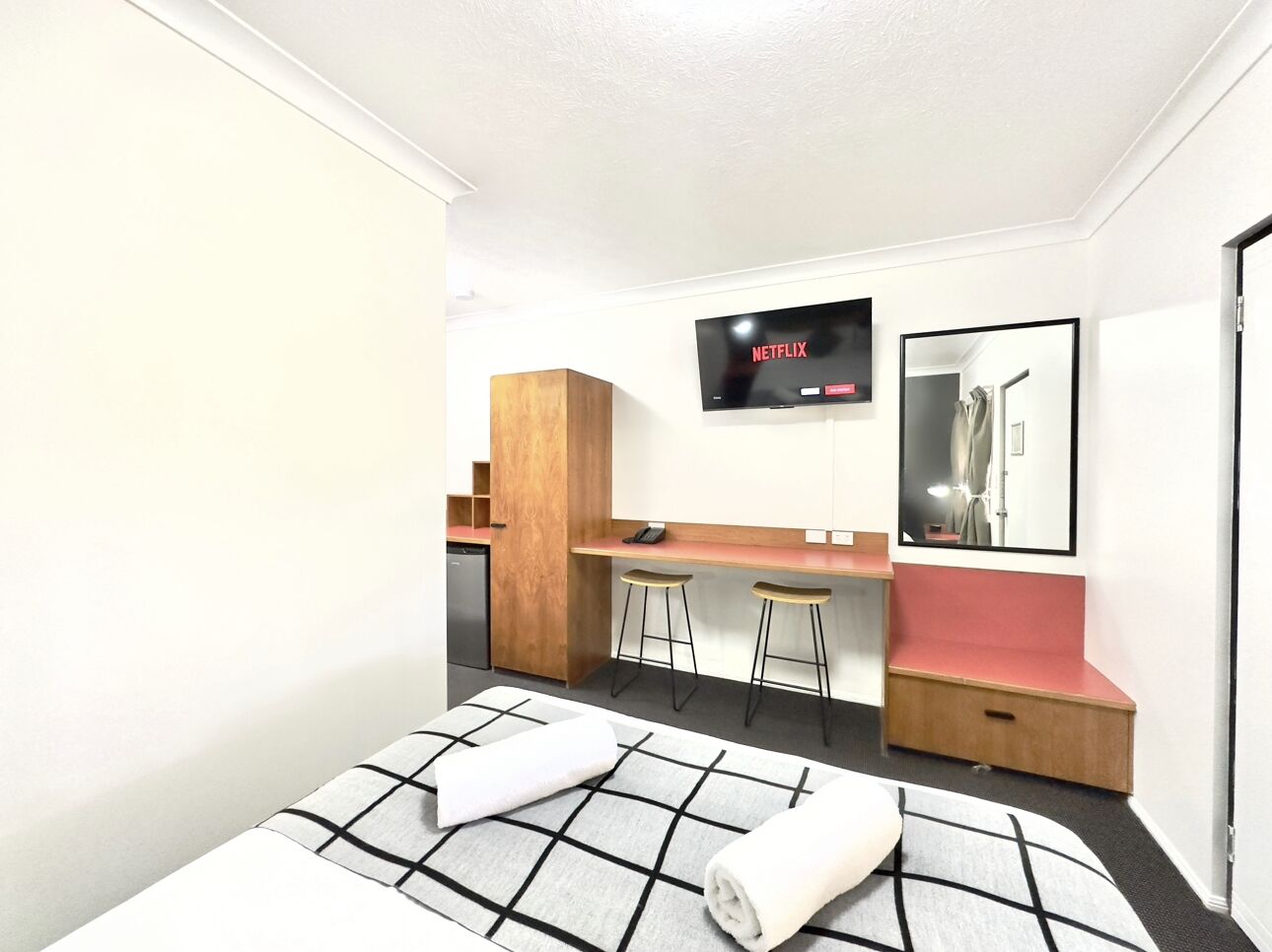 A modern hotel room with a bed, a desk, two stools, a television, and a mirror.
