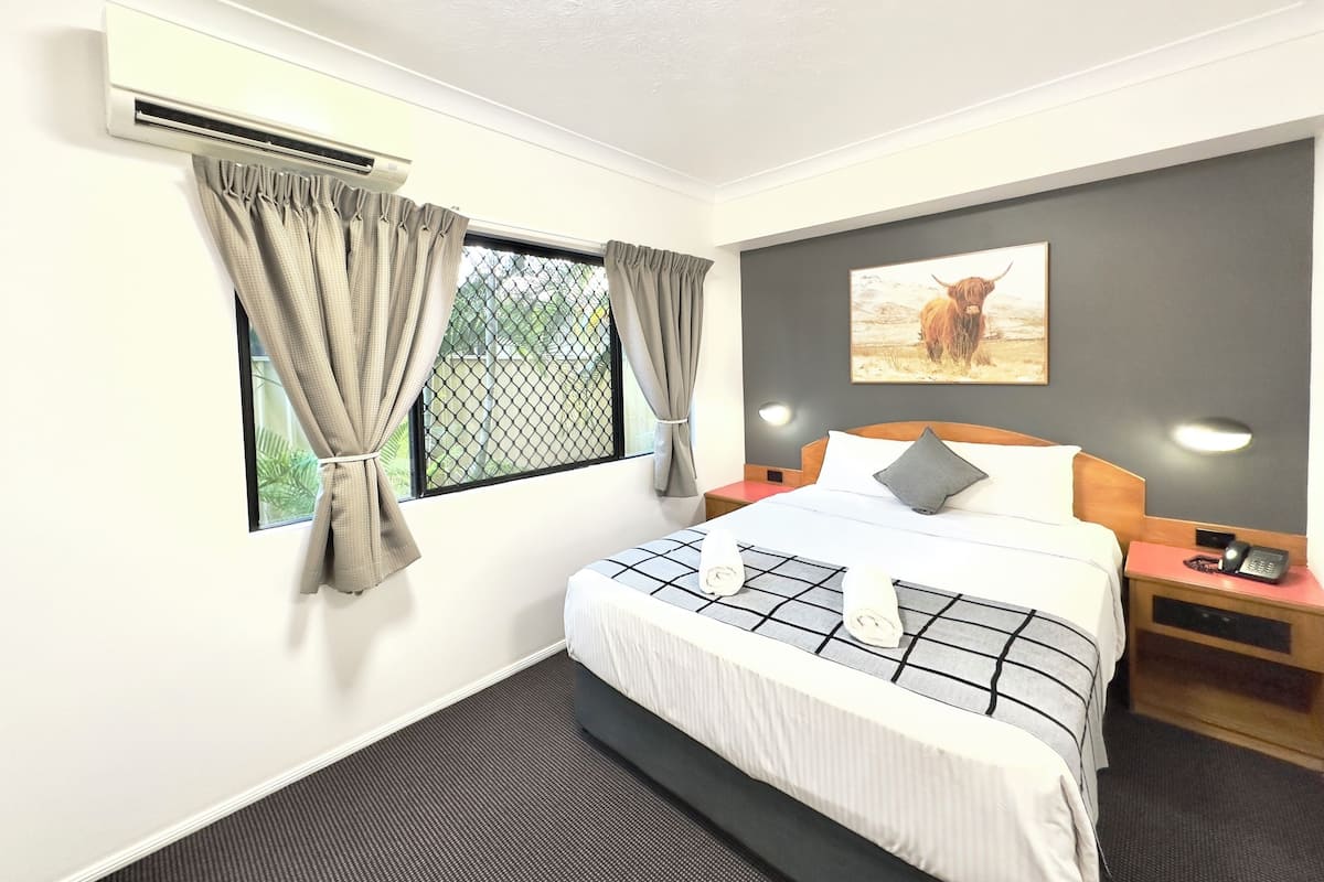 A hotel room with a bed, a nightstand, a wall-mounted phone, and a window with curtains.