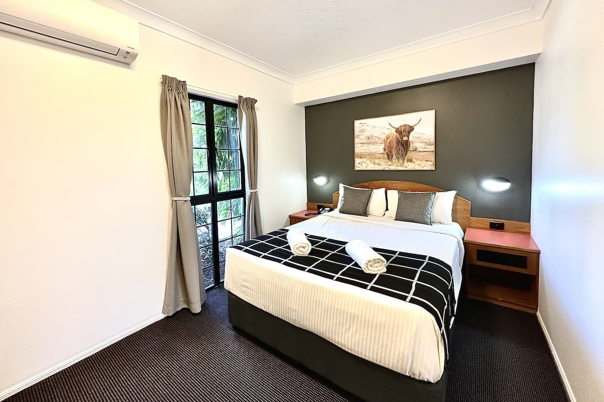 A hotel room with a large bed, a window with curtains, a wall-mounted artwork of a cow, and a bedside table with a lamp.