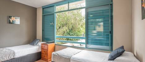 A bedroom with two beds, a window with blue shutters, a small wooden table, and a wall-mounted shelf.