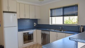 Una cocina con armarios blancos, un refrigerador de acero inoxidable, un horno, un lavavajillas, un fregadero y una ventana con persianas.