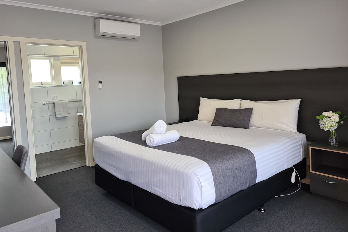 A modern hotel room with a large bed, a bedside table, a nightstand, a desk, and a bathroom with a mirror and towel rack.