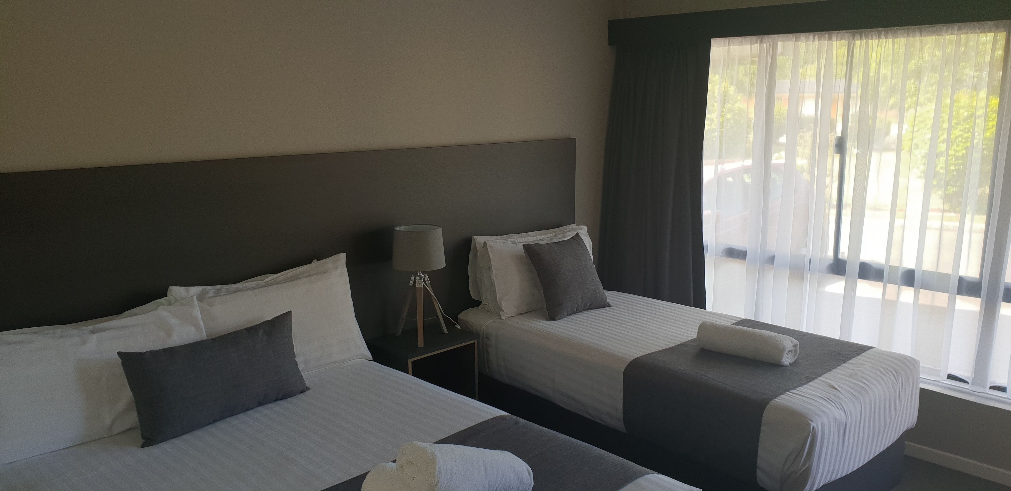Two single beds with white and gray bedding, a bedside lamp, and a window with sheer curtains.