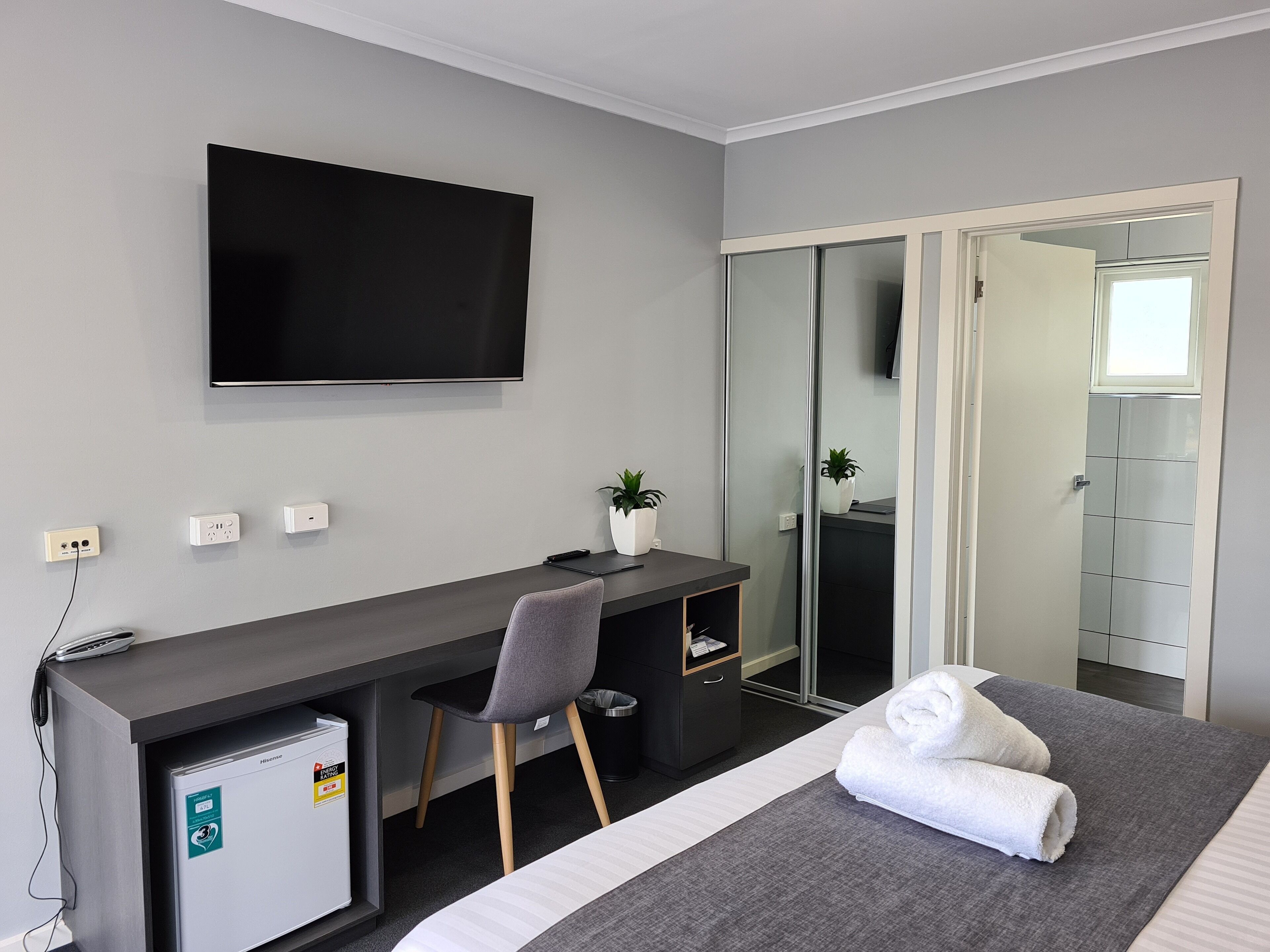 A hotel room with a bed, a desk with a chair, a TV mounted on the wall, a mini-fridge, and a bathroom with a mirror and toilet.
