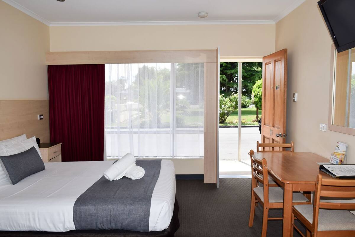 A hotel room with a bed, a desk, a chair, a window with a view of greenery, and a door leading to an outdoor area.