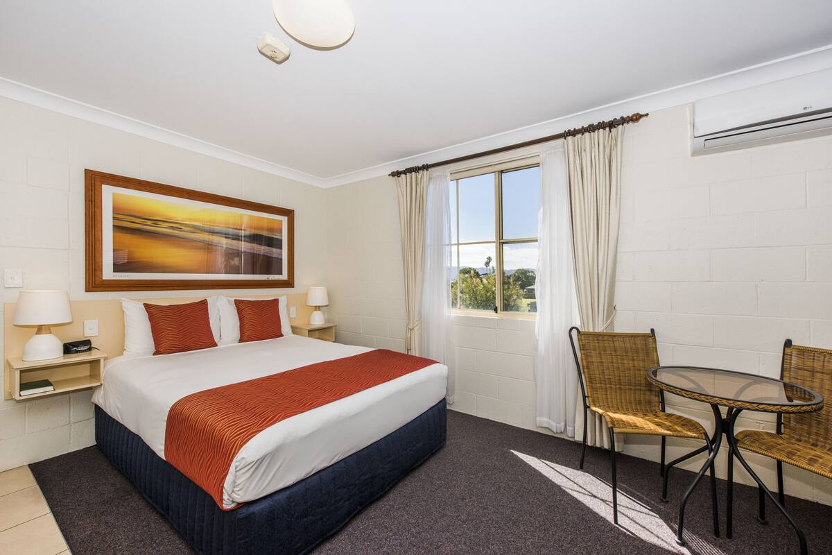 A hotel room with a bed, two chairs, a table, a window with curtains, a painting on the wall, and an air conditioning unit.
