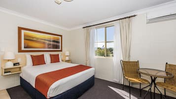 A hotel room with a bed, two chairs, a table, a window with curtains, a painting on the wall, and an air conditioning unit.