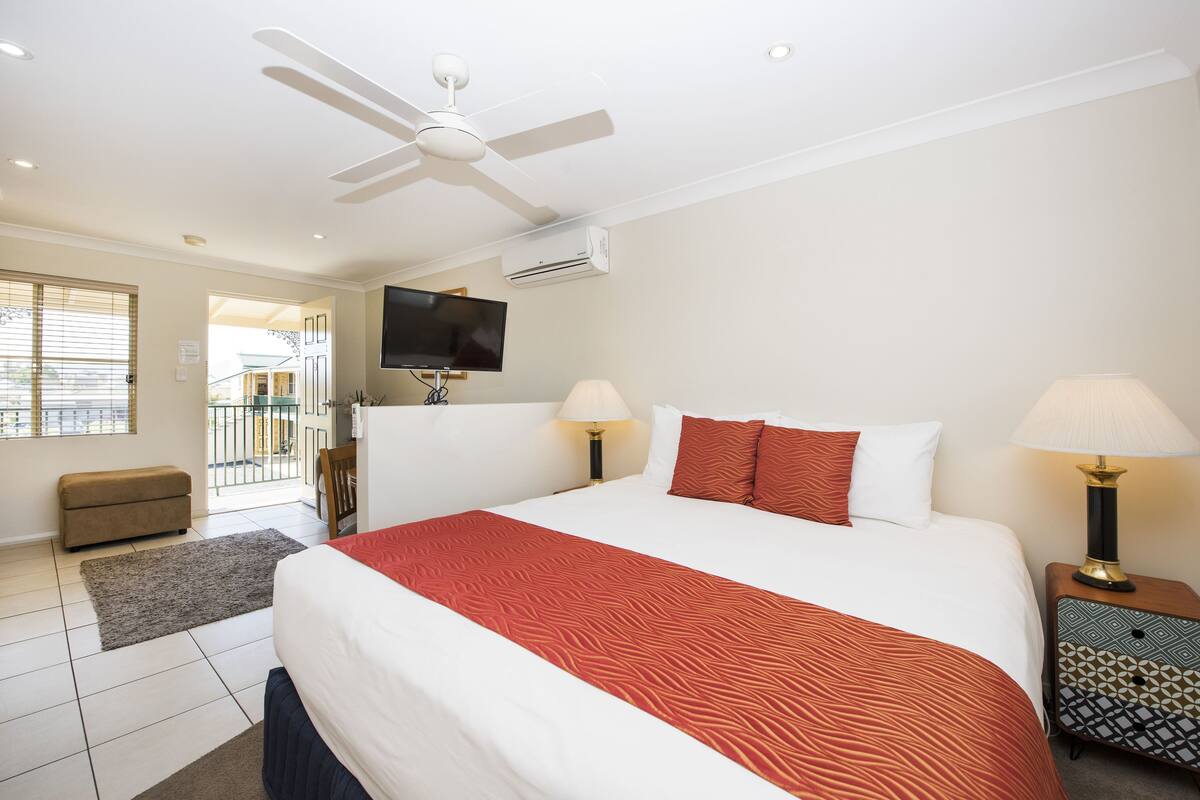 A hotel room with a large bed, two bedside lamps, a ceiling fan, an air conditioning unit, a flat-screen TV, and a small sofa.
