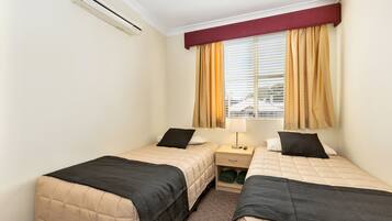A hotel room with two beds, a window with curtains, a bedside table with a lamp, and an air conditioning unit.