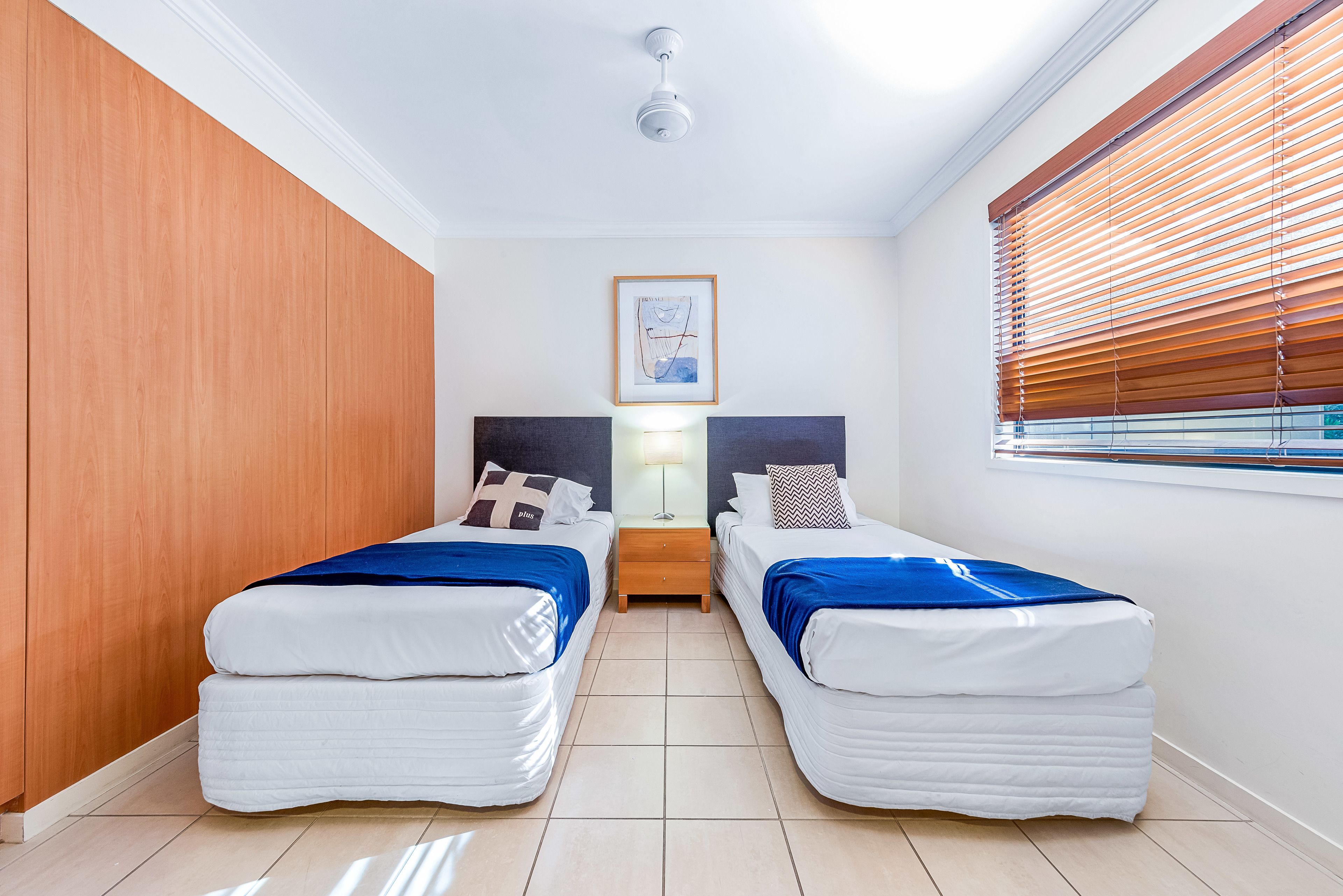 A bedroom with two beds, a nightstand, a ceiling fan, and a window with blinds.