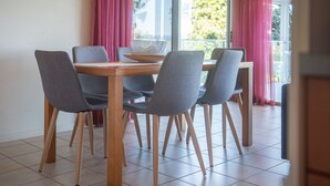 Comedor con mesa de madera, sillas grises, cortinas rosadas y vista a zonas verdes.