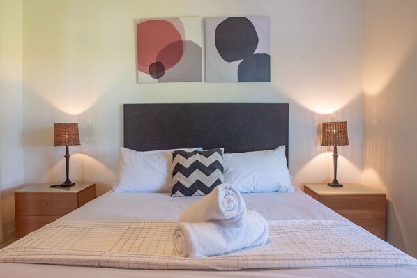 A neatly made bed with a rolled towel, two nightstands with lamps, a black and white pillow, and abstract wall art. - Oasis 22 (Whitsundays)