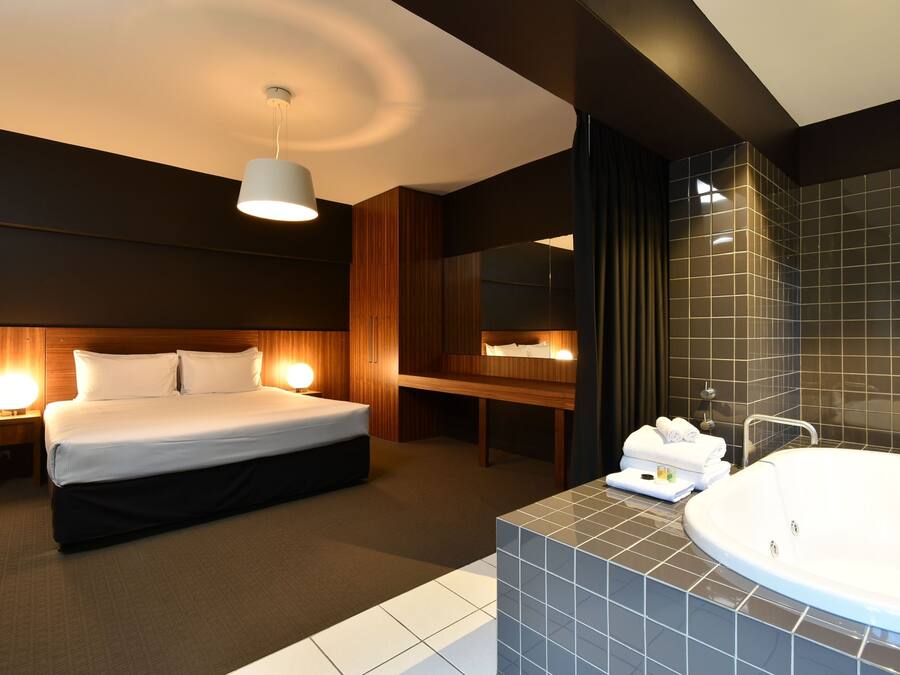 A modern hotel room with a large bed, bedside lamps, a vanity with a bathtub, and a mirror.