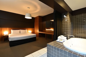 A modern hotel room with a large bed, bedside lamps, a vanity with a bathtub, and a mirror.