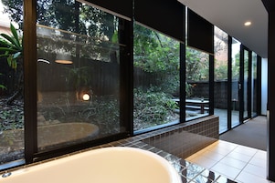 A modern bathroom with a large bathtub, a glass enclosure with a view of a garden, and a tiled floor.