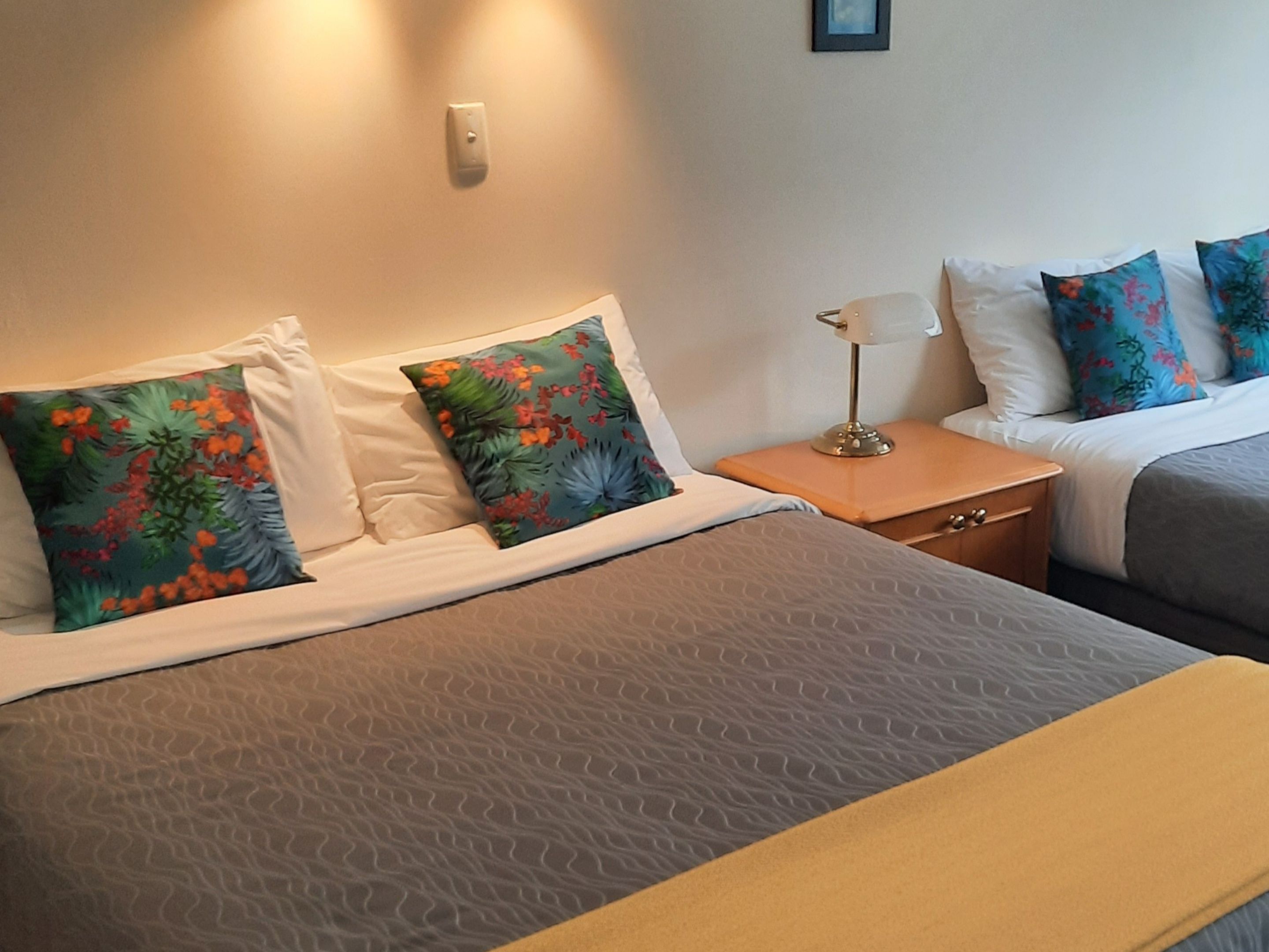 Habitación de hotel con dos camas, cada una con un edredón de estampado diferente. Entre las camas, un mesita de noche con una lámpara, y una lámpara de pared sobre la mesita.