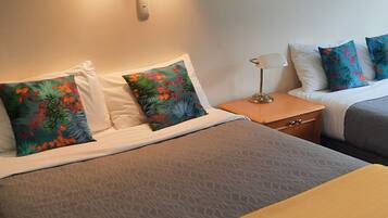 Habitación de hotel con dos camas, cada una con un edredón de estampado diferente. Entre las camas, un mesita de noche con una lámpara, y una lámpara de pared sobre la mesita.