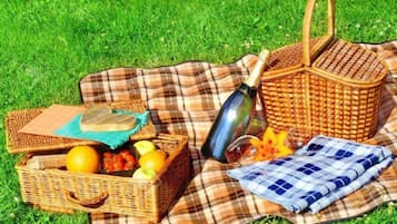 Un montaje para picnic en un área cubierta de césped, con una cesta de mimbre, un recipiente metálico, un paño y una manta a cuadros.