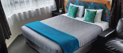 A bedroom with a bed, a headboard, a window with curtains, a small table, and a blue rug.