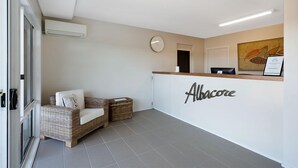 A reception area with a wicker sofa, a reception desk with 'Albacore' signage, a clock, and a framed artwork on the wall. - Albacore Apartments (Merimbula)