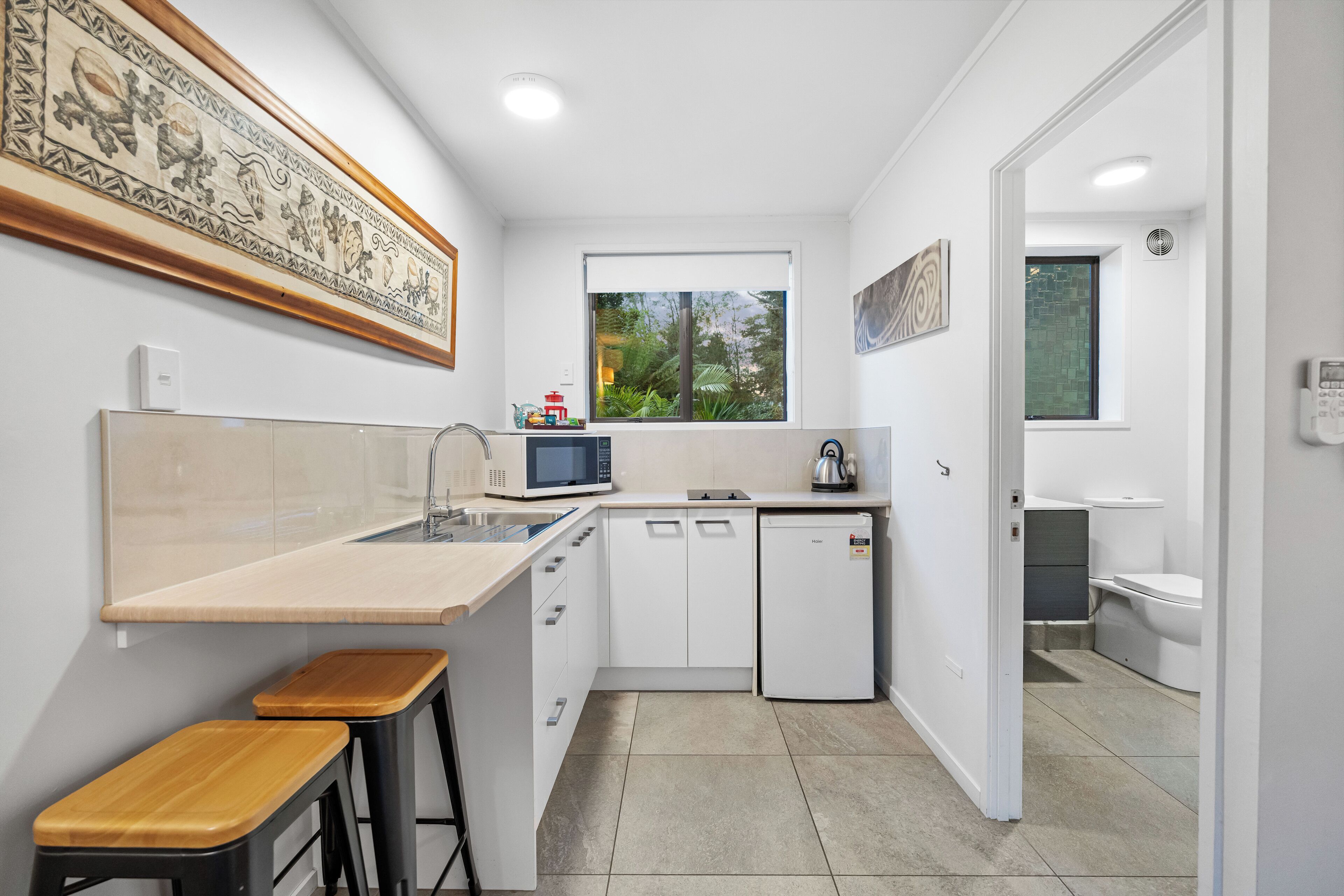 Cocina compacta con microondas, lavamanos y ventana. Hay dos taburetes y un refrigerador pequeño.