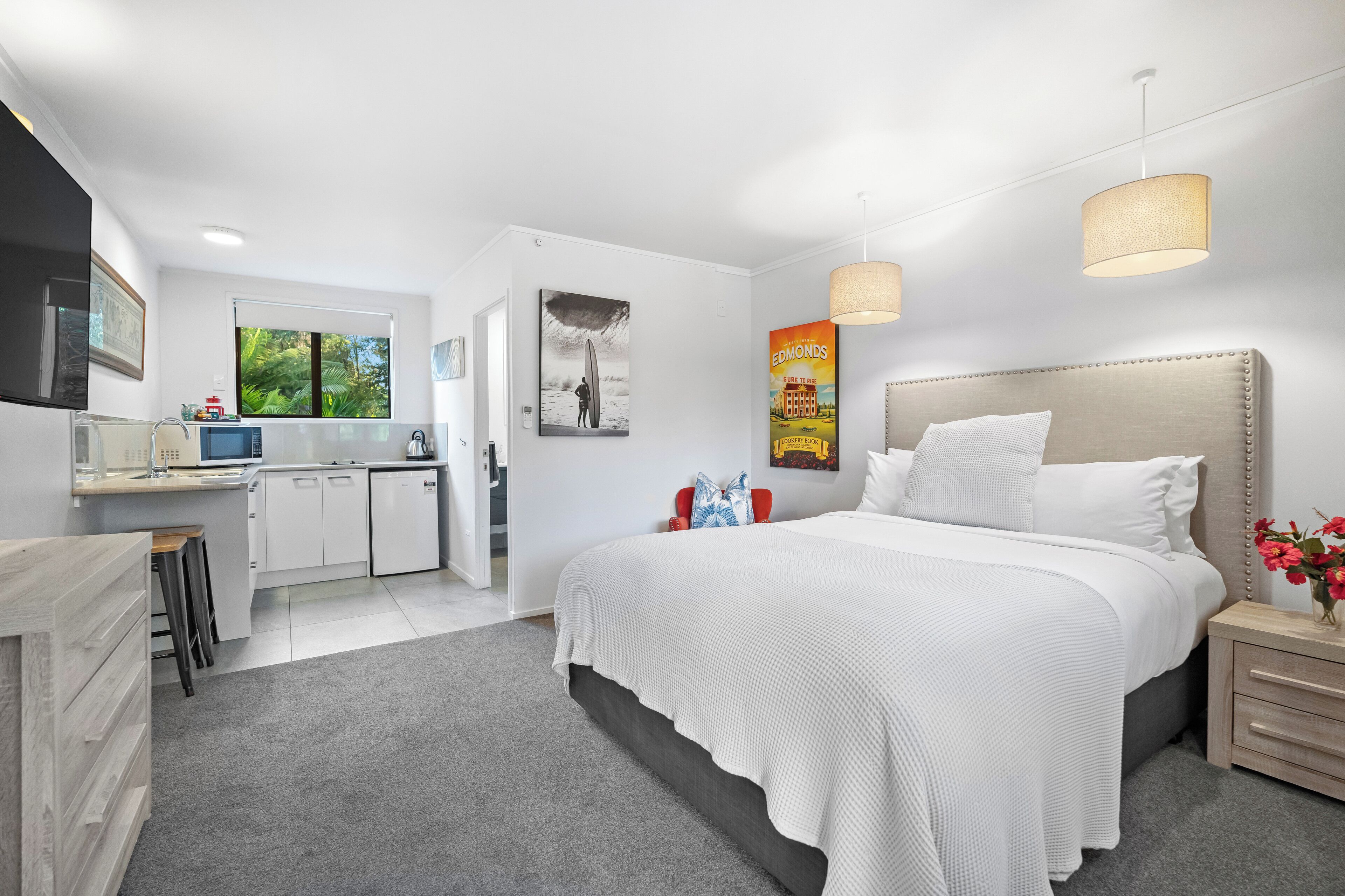 A modern bedroom with a large bed, bedside tables, a desk, and a kitchen area.