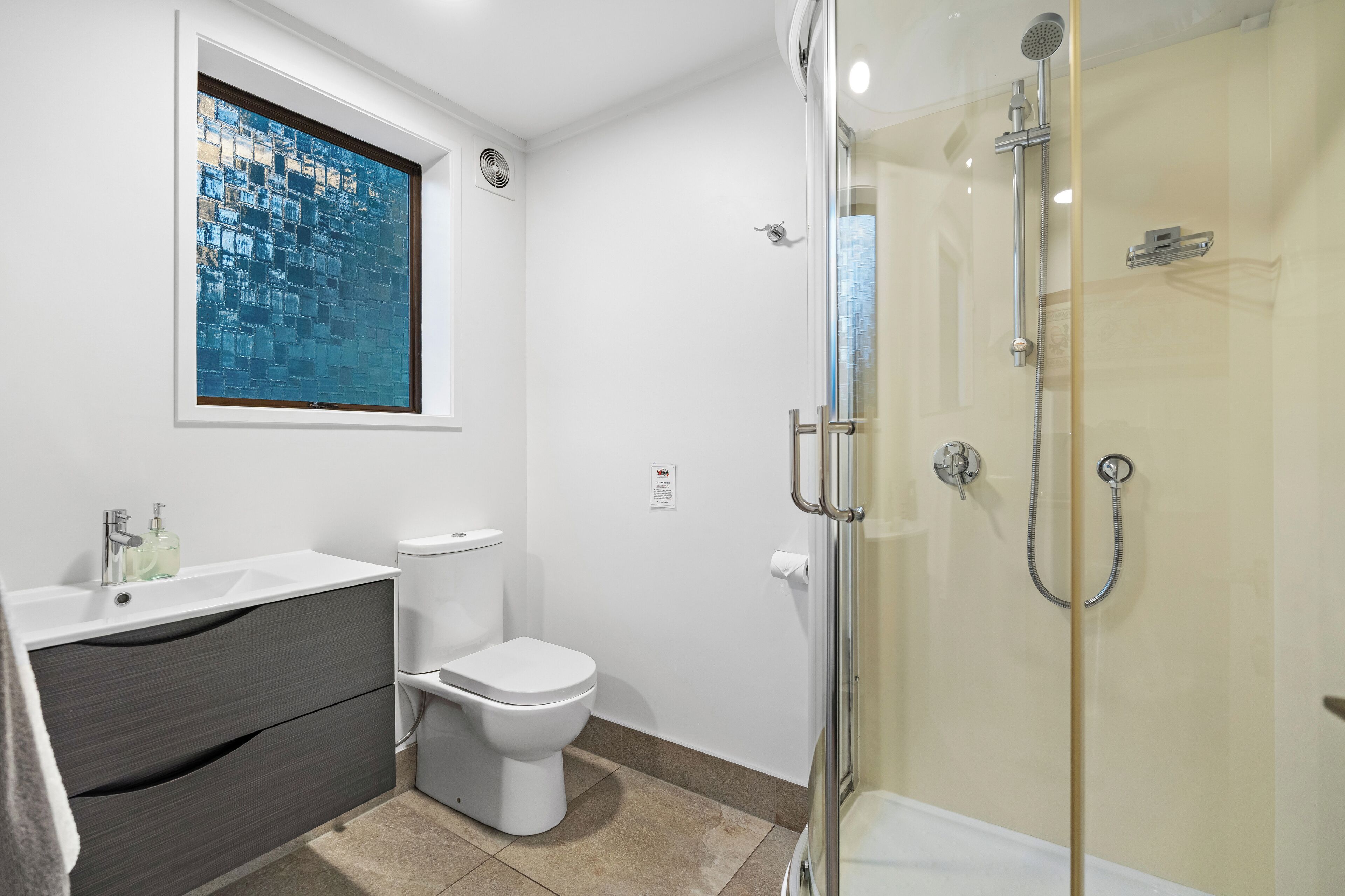 Et badeværelse med en brusekabine i glas, et hvidt toilet, et mørkegråt toiletbord og et vindue med udsigt til et blåt mosaikmønster.