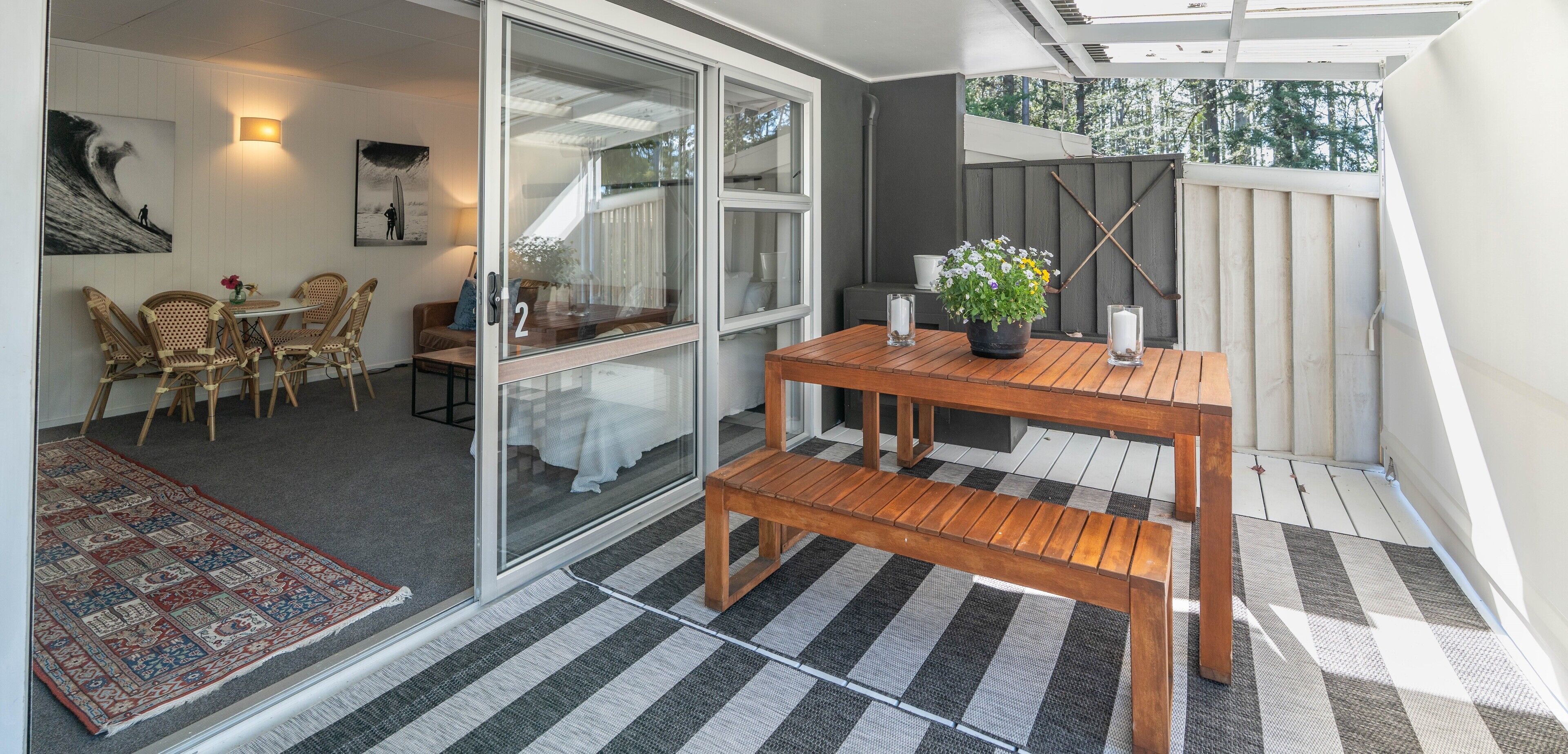A balcony with a wooden table, two chairs, a potted plant, and a rug.