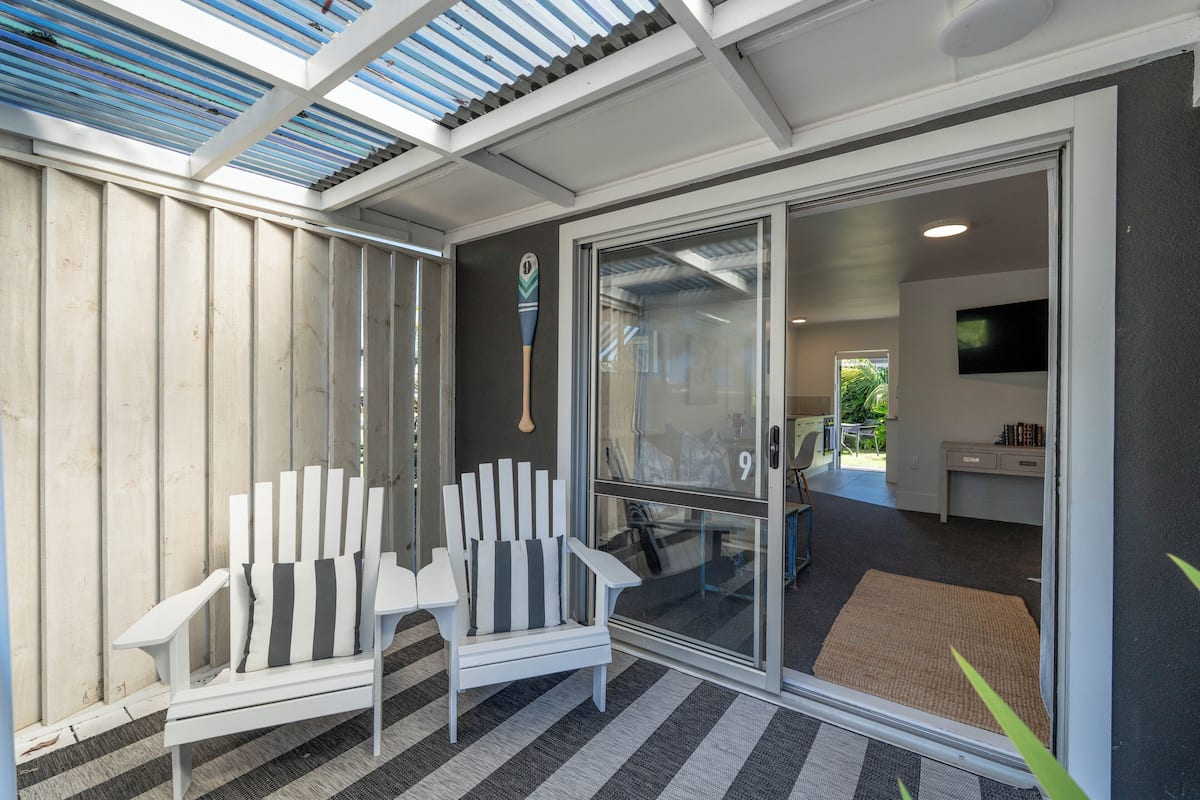 A modern room with a glass door leading to a patio area, a white chair with a striped cushion, and a small desk with a chair.