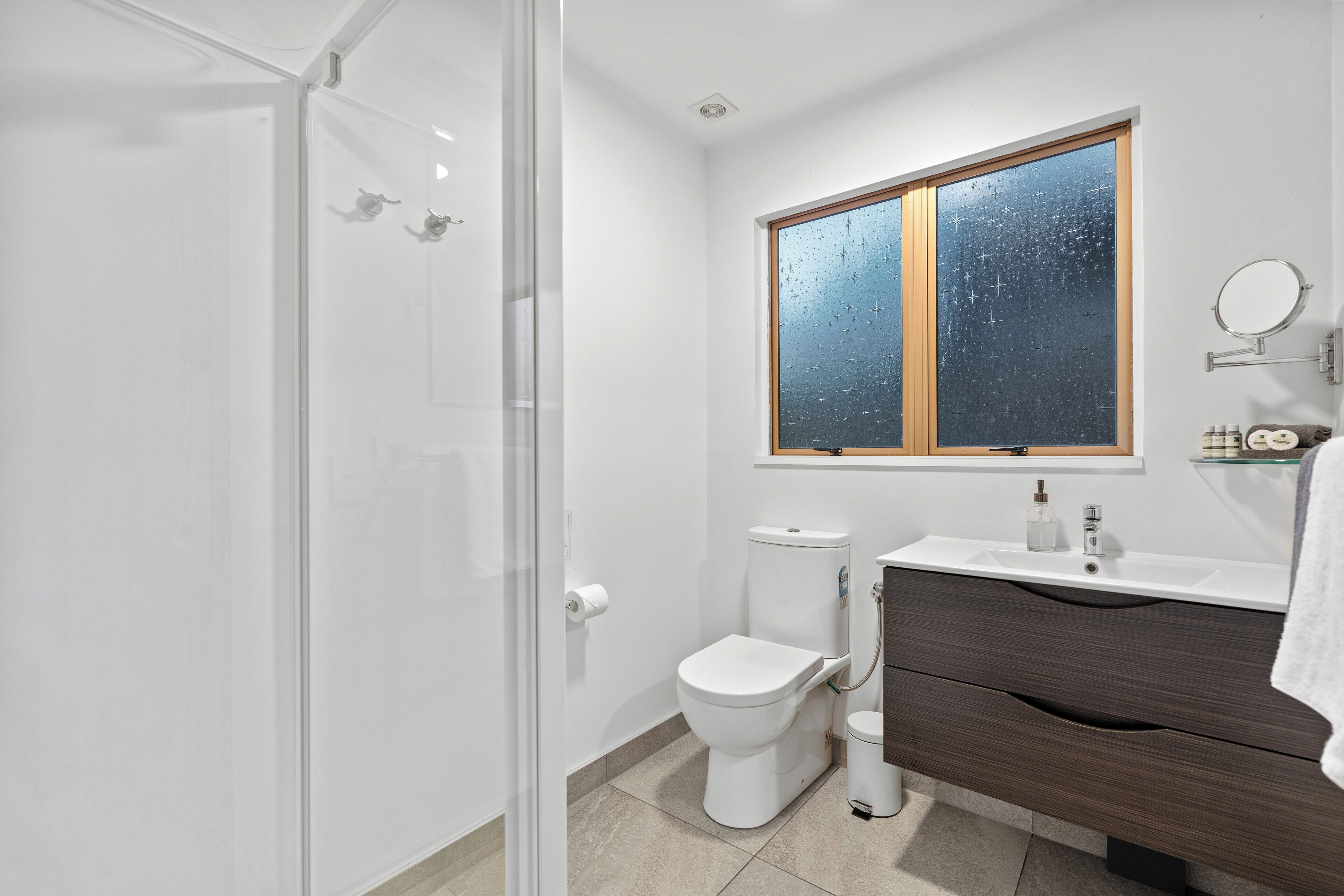 Un baño moderno con cabina de ducha de vidrio, inodoro blanco, tocador con espejo y ventana con vistas a las estrellas.