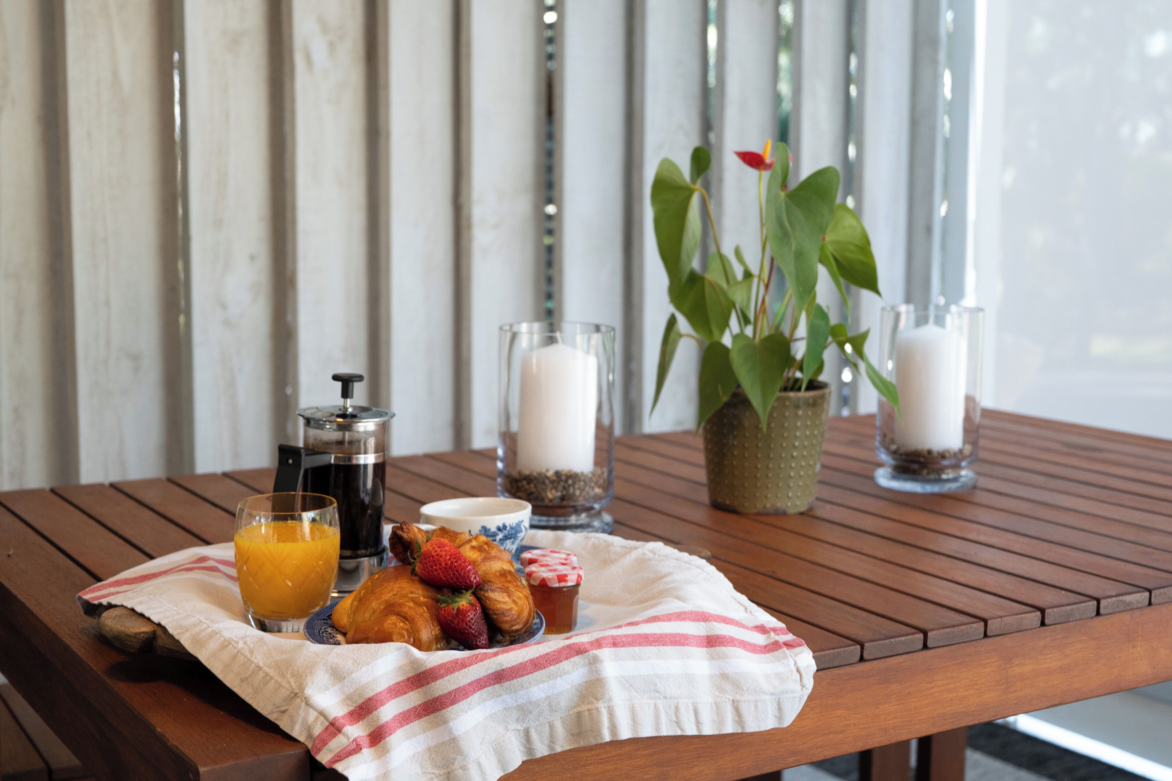 A wooden table with breakfast items including croissants, strawberries, orange juice, and coffee. There are candles and a potted plant on the table.