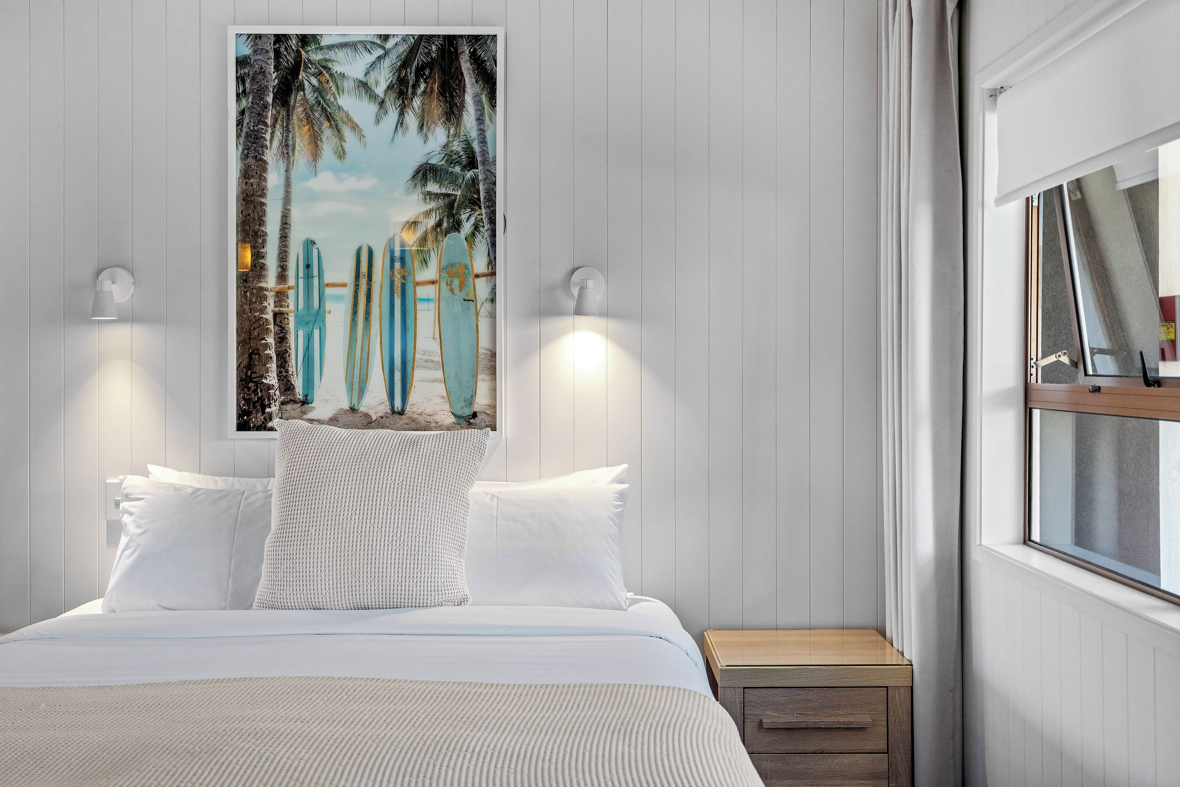 A bedroom with a bed, white bedding, a nightstand, a lamp, and a beach-themed painting on the wall.