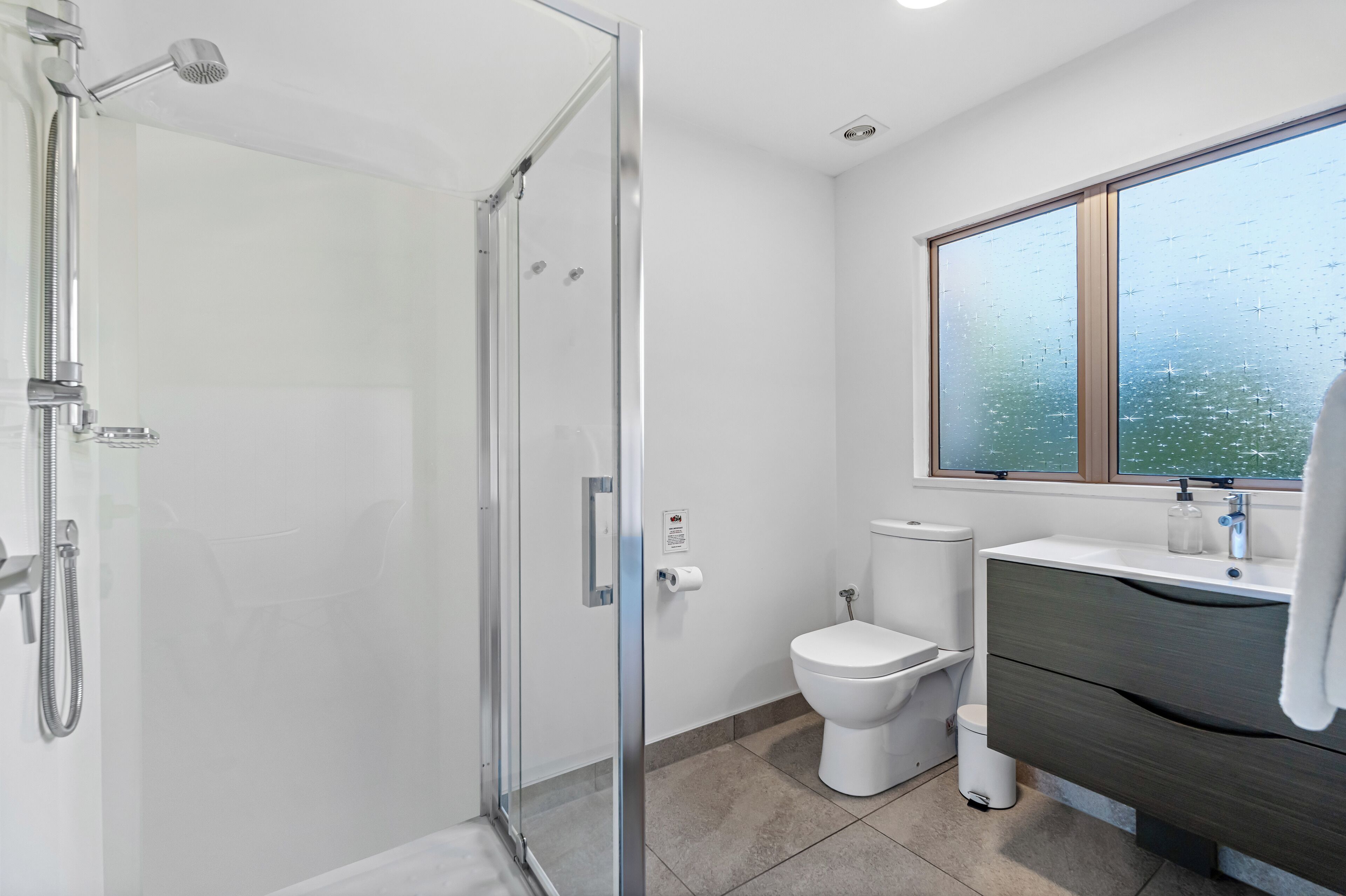 A modern bathroom with a glass-enclosed shower, a toilet, and a vanity with a large window above.