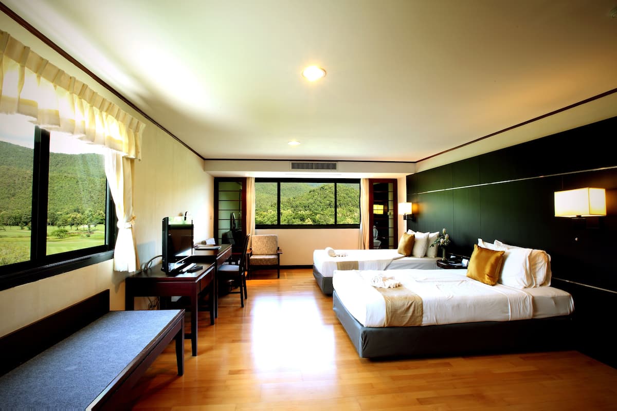 A hotel room with two beds, a desk, a chair, and a view of a green landscape through large windows.