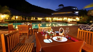 A dining area with a table set for two, a pool, and a building in the background. - Alpine Golf Resort Chiang Mai (Ban Thi)