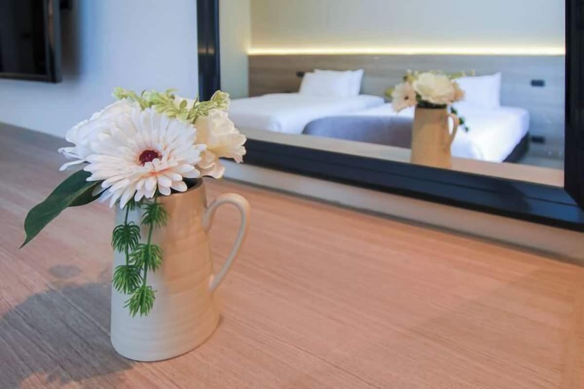 A white vase with flowers on a wooden surface, a bed with white bedding, and a mirror reflecting the room.