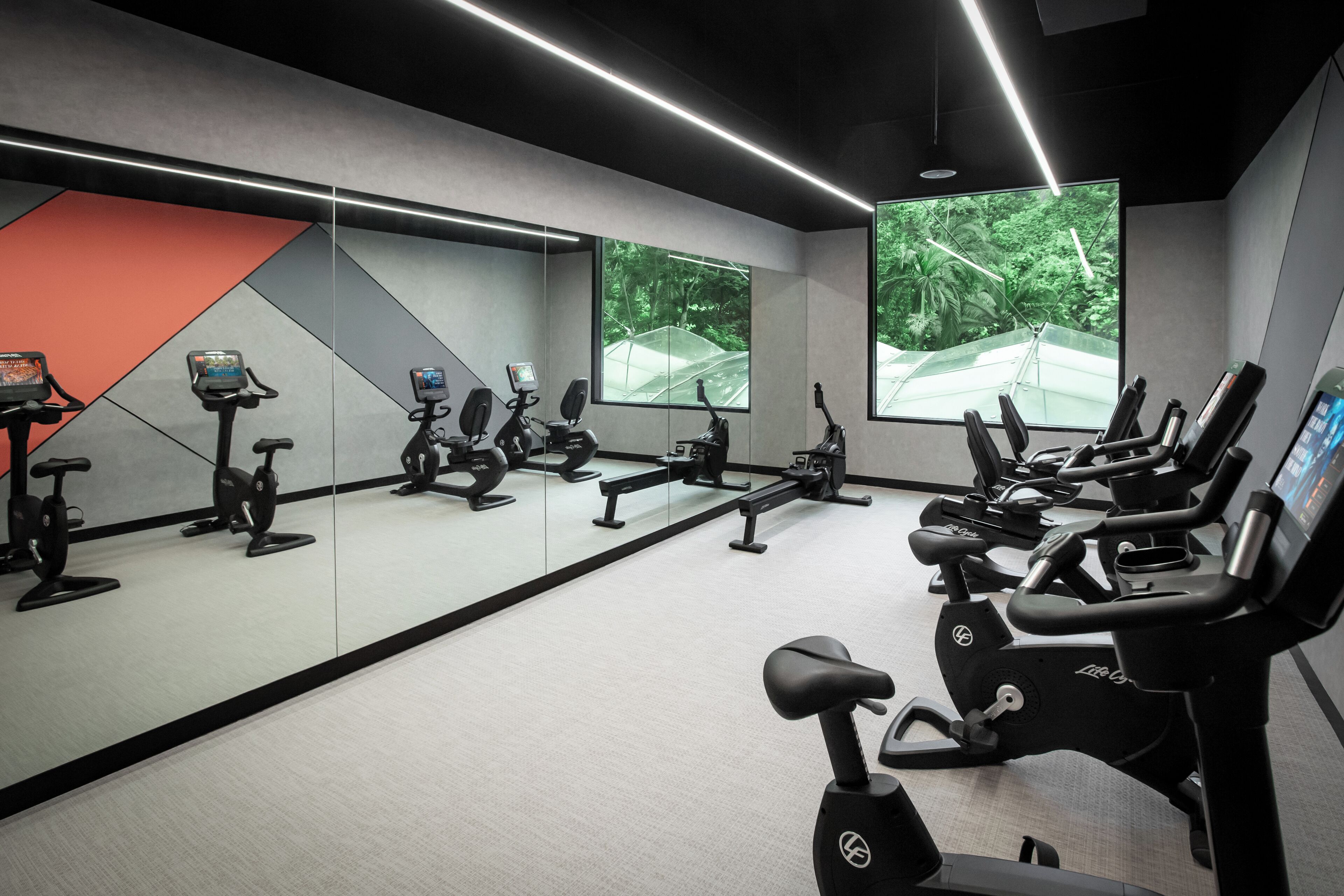 Un gimnasio con bicicletas estáticas, espejos y amplias ventanas con vistas a la vegetación.