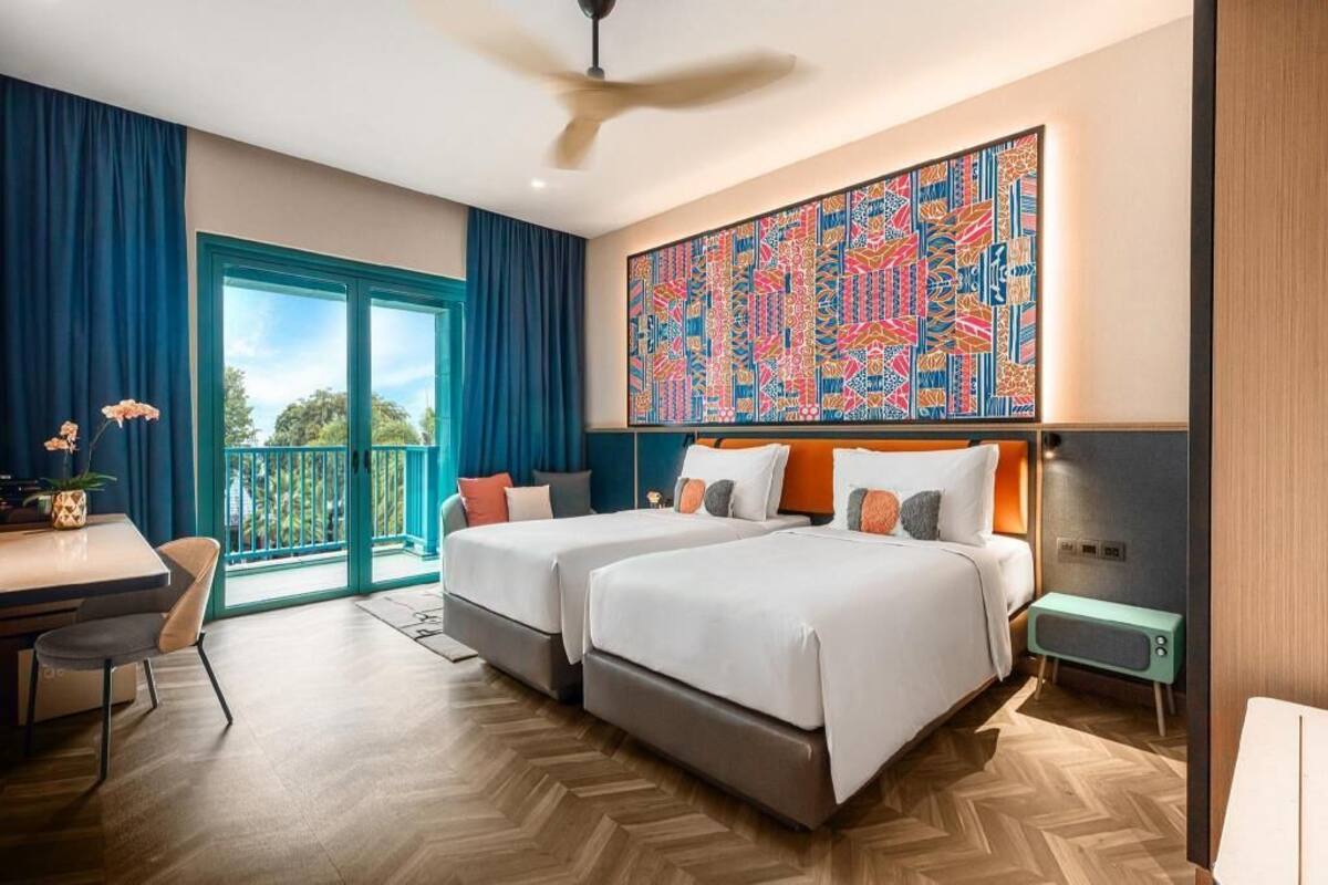 Une chambre d’hôtel avec deux lits, un bureau, une chaise, un balcon avec des rideaux bleus et une peinture abstraite aux couleurs vives accrochée au mur.