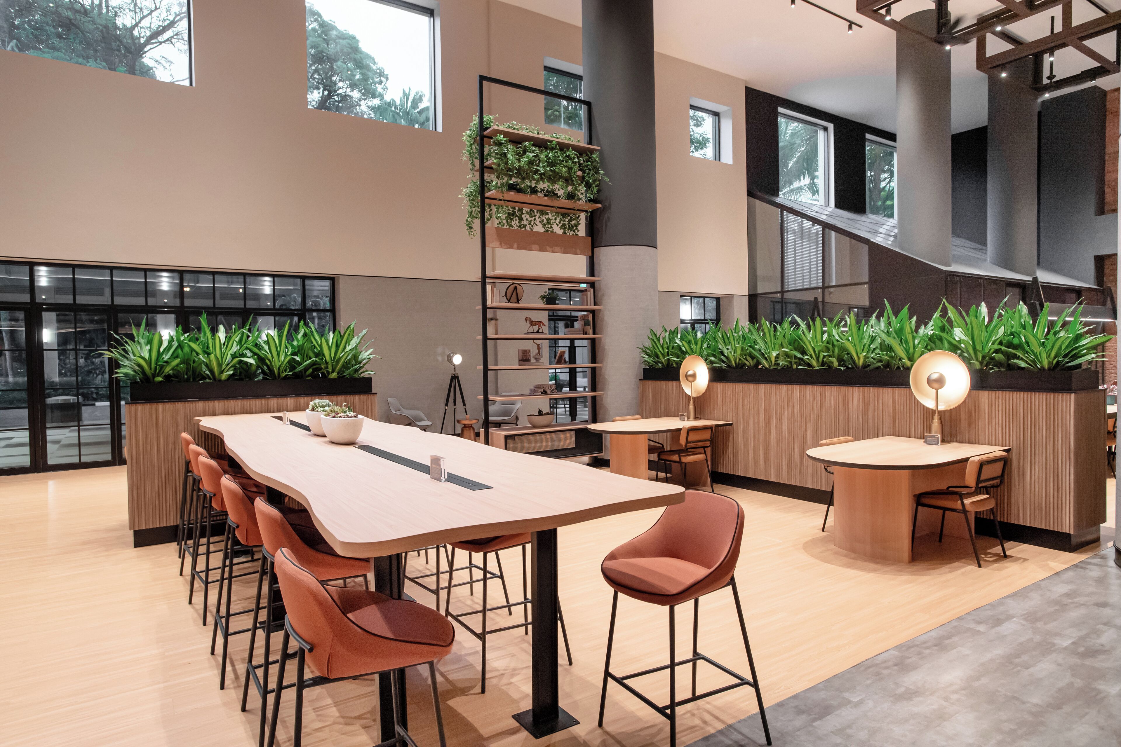 A modern interior with a long communal table, several chairs, a bookshelf with plants, and a staircase.