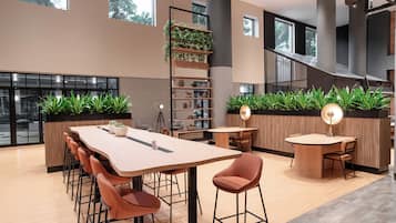 A modern interior with a long communal table, several chairs, a bookshelf with plants, and a staircase.