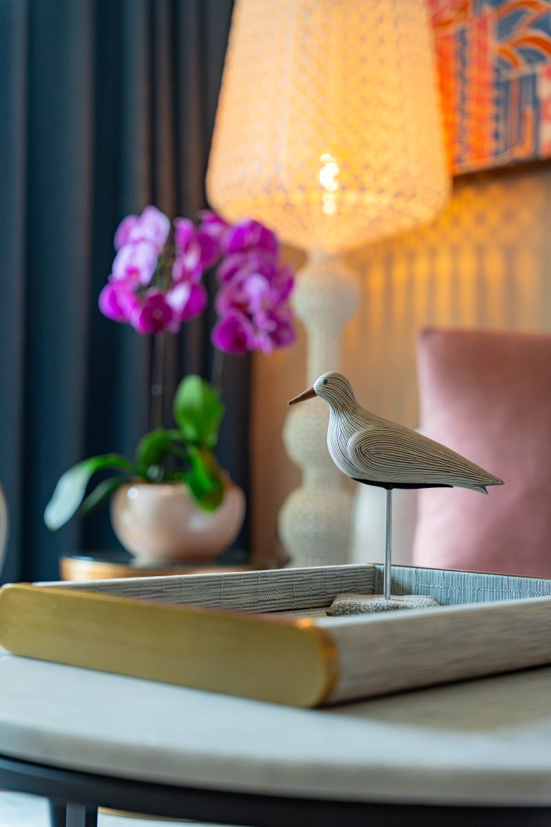 A bird figurine on a book, a lamp, and purple flowers in the background.