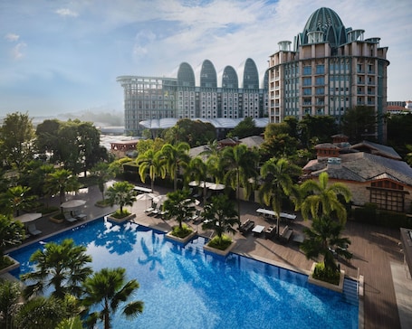 A hotel with a large pool, palm trees, and a modern building with unique architecture in the background.