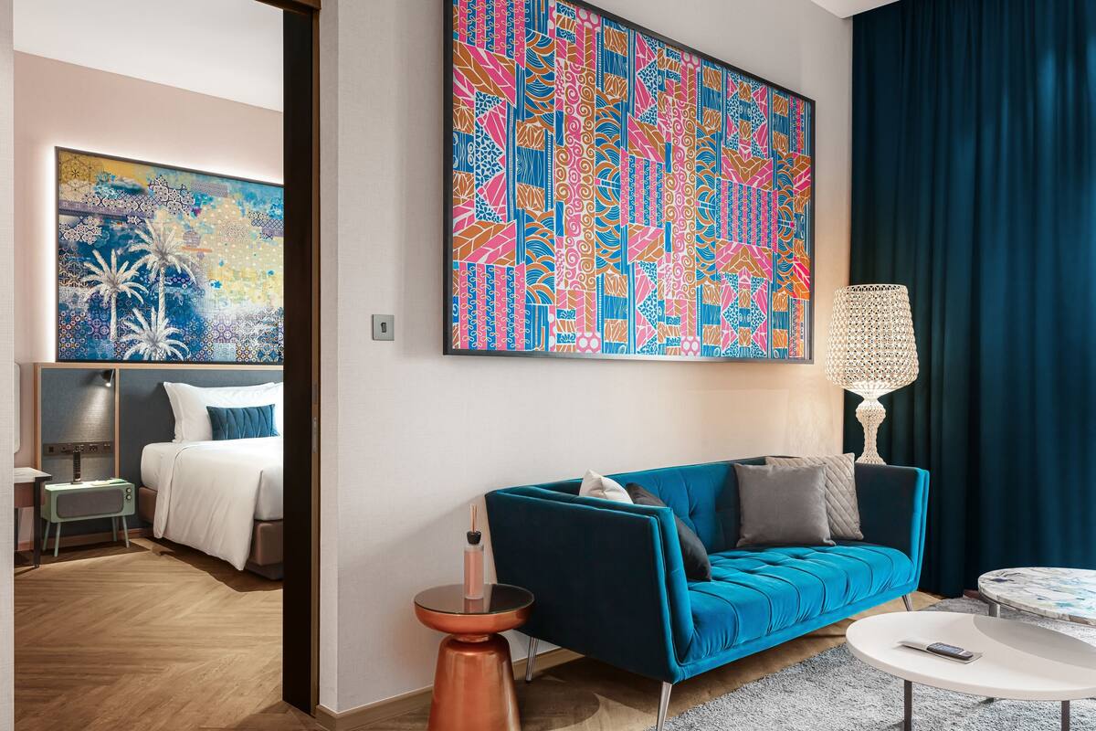 Une chambre d’hôtel moderne avec un canapé bleu, une table basse ronde et un grand tableau abstrait accroché au mur.